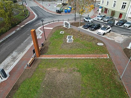 Dreieckiger Grünbereich mit einem schmalen Weg und einer hohen orangefarbenen Skulptur an einer Straßenecke mit geparkten Autos