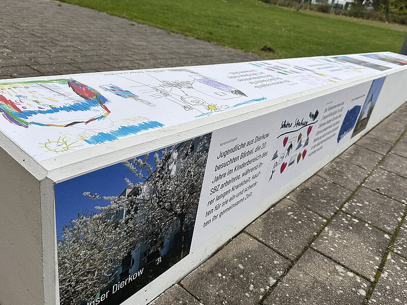 Lange Sitzbank mit Fotos, Texten und bunten Kinderzeichnungen auf der Oberseite, auf gepflastertem Boden im Freien