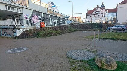 Freifläche mit zwei runden Gullydeckeln, einem großen Stein und einer mit Graffiti bedeckten Mauer im Hintergrund