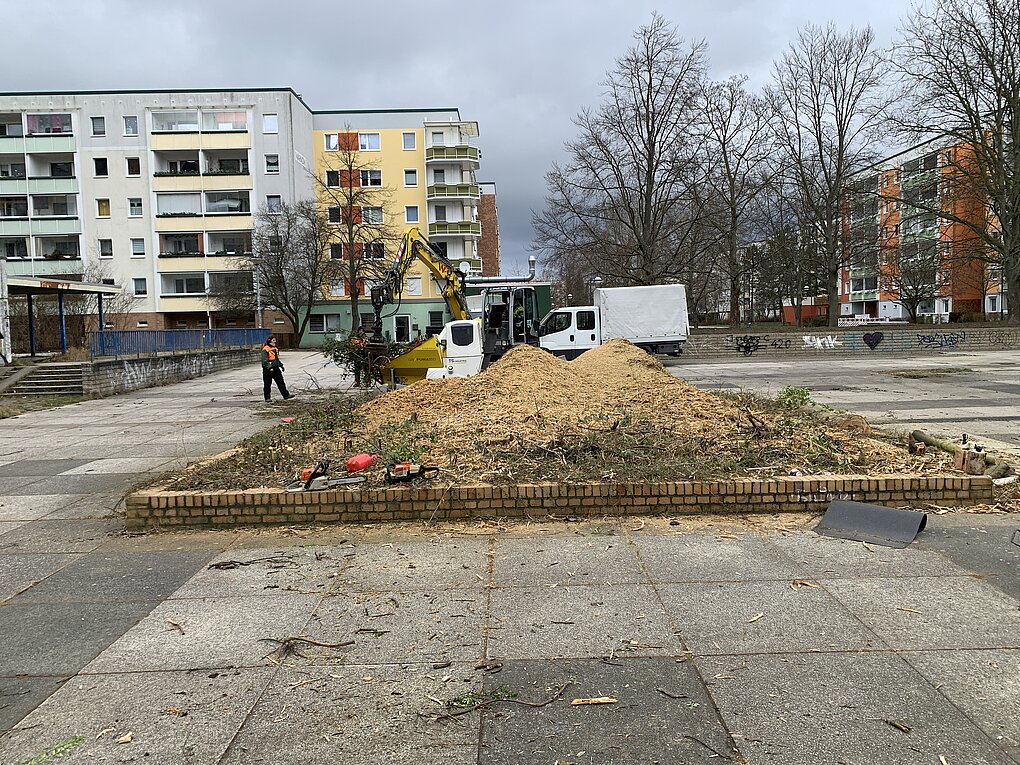Baustelle mit Erdbewegungen und Baumstämmen vor mehrstöckigen Wohngebäuden im Hintergrund.