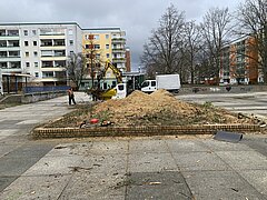 Baustelle mit Erdbewegungen und Baumstämmen vor mehrstöckigen Wohngebäuden im Hintergrund.