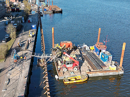 Bauarbeiten an einem Hafenbecken mit einer Arbeitsplattform auf dem Wasser, auf der Baumaschinen und Baumaterialien liegen, neben einer Uferpromenade mit Bauarbeitern und Fahrzeugen