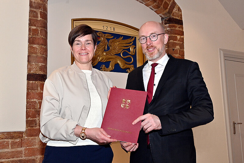 Zwei Personen in formeller Kleidung übergeben eine rote Urkunde vor einer Wand mit einem Wappen und einer Ziegelstein-Bogennische