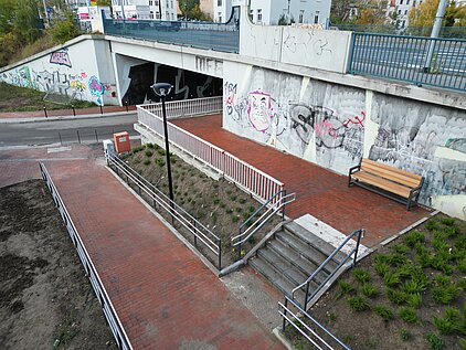 Luftaufnahme eines mit roten Ziegeln gepflasterten Wegs mit Treppe und Rampe neben einer Unterführung mit Graffiti an den Wänden und einer Holzbank