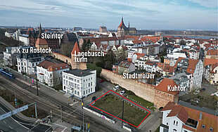 Luftaufnahme von Rostock mit beschrifteten Sehenswürdigkeiten: Steintor, Lagebuschturm, Stadtmauer und Kuhtor. Im Vordergrund das freie Grundstück
