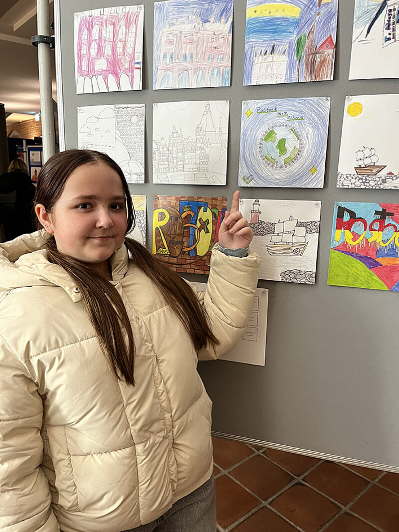 Person in hellbeiger Steppjacke zeigt mit ausgestrecktem Zeigefinger auf eine Wand mit mehreren bunten und schwarz-weißen Kinderzeichnungen auf ihr Bild mit einer Weltkugel