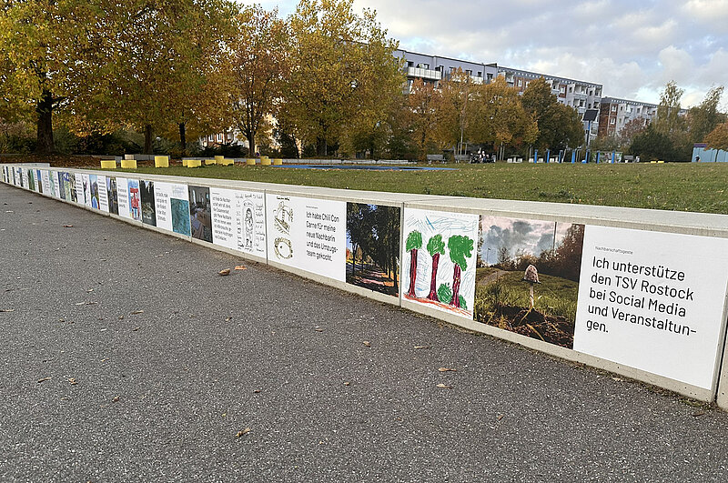 Lange Sitzbank mit einer Reihe von quadratischen Bildern und Texten, die an der Vorderseite angebracht sind, im Freien vor herbstlich gefärbten Bäumen und Wohngebäuden