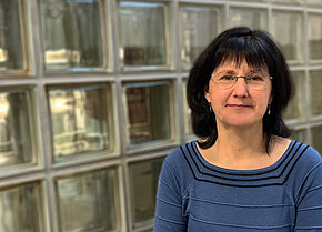 Frau mit schulterlangen Haaren und Brille, trägt ein blaues Oberteil mit Streifen, steht vor einer Glaswand..