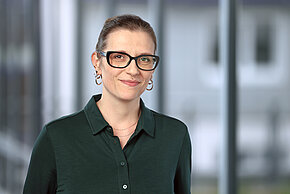 Person mit zusammengebundenem Haar, Brille, Ohrringen und dunkelgrünem Hemd vor unscharfem Bürohintergrund.