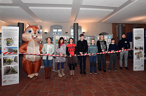 Mehrere Personen stehen in einer Reihe hinter einem rot-weiß gestreiften Absperrband in einem Raum mit Backsteinboden und Informationsständen, links eine Person im Eichhörnchen-Kostüm