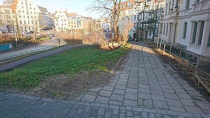 Gepflasterter Gehweg neben einem grasbewachsenen Hang mit Büschen und mehrstöckigen Wohngebäuden im Hintergrund