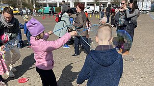 Kinder und Erwachsene machen Seifenblasen auf einem sonnigen Platz vor einem Wohnhaus