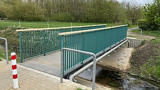Grüne Fußgängerbrücke in Lichtenhagen über einen kleinen Bach, umgeben von Wiese und Bäumen. Geländer aus Metall, Zugang über eine Rampe.