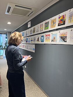 Frau mit dunkler Kleidung steht seitlich vor einer Wand mit zahlreichen farbigen Bildern in einem Flur.