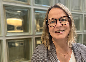 Porträt einer lächelnden Frau mit Brille. Sie trägt einen Blazer und hat schulterlanges, blondes Haar.