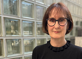 Porträt einer Frau mit schulterlangen Haaren und Brille, die in einem schwarzen Pullover mit Rüschendetails gekleidet ist. Sie steht vor einer Glasbauwand.en.