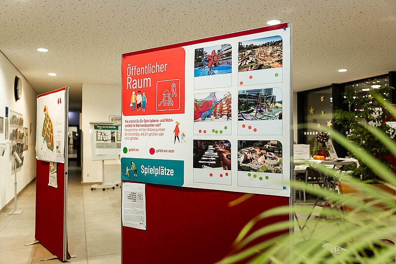 Informationswand mit mehreren farbigen Postern und Texten in einem Innenraum mit Pflanzen im Hintergrund