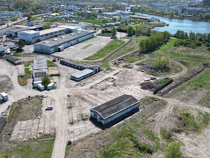 Luftaufnahme des unbebauten Gebietes mit mehreren Lagerhallen, unbefestigten Flächen und der Unterwarnow  im Hintergrund