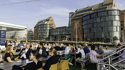 Blick auf eine belebte Terrasse auf einem Schiff mit Gästen, umgeben von modernen Gebäuden und einem klaren Himmel.