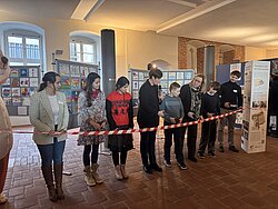 Gruppe von Menschen steht in einer Reihe in einem Raum mit hohen Fenstern und hält ein langes rotes Band