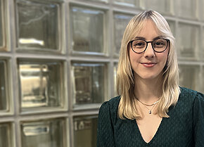 Junge Frau mit blonden, langen Haaren und Brille, trägt ein grünes Oberteil, lächelt vor einer Wand aus Glasbausteinen.