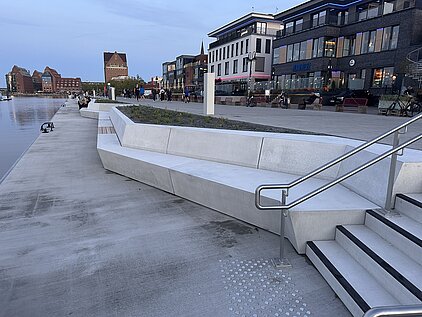Lange, moderne Sitzbank aus Beton an einem Ufer mit Treppe und Geländer im Vordergrund und mehrstöckigen Gebäuden im Hintergrund