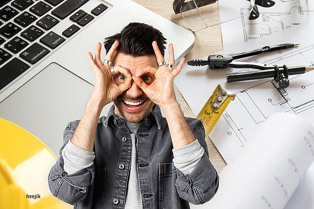 Junger Mann mit fröhlichem Gesichtsausdruck hält seine Hände in Form von Brillen vor die Augen, umgeben von einem Laptop und technischen Zeichnungen.