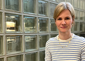 Frau mit blonden Haaren und blauer gestreifter Bluse, lächelt vor einer Wand aus Glasbausteinen.
