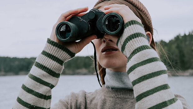 Frau mit gestreiftem Pullover und Schal, die mit einem Fernglas auf einen See blickt. Im Hintergrund sind Bäume und ein bewölkter Himmel zu sehen.