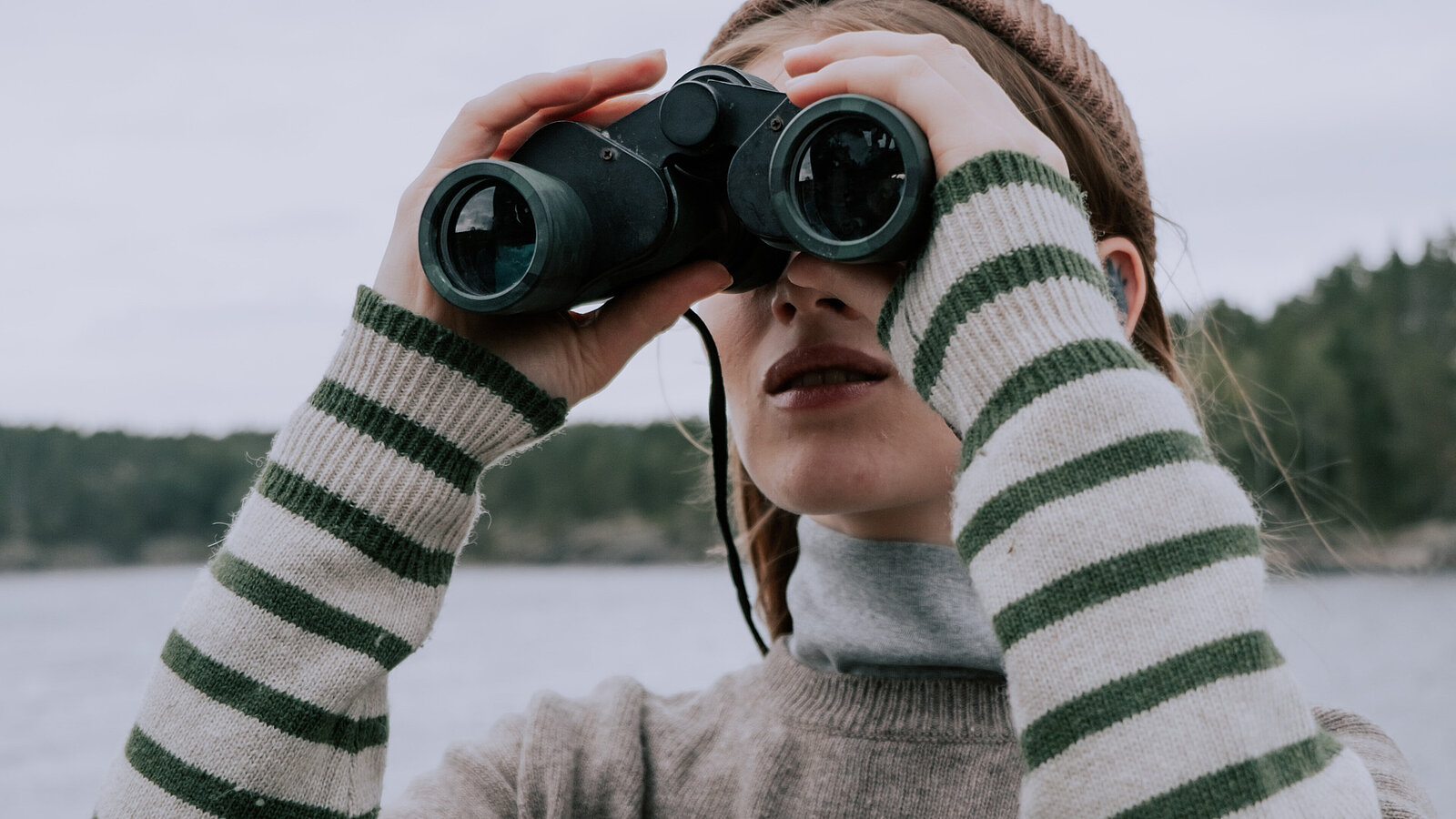 Frau mit gestreiftem Pullover und Schal, die mit einem Fernglas auf einen See blickt. Im Hintergrund sind Bäume und ein bewölkter Himmel zu sehen.