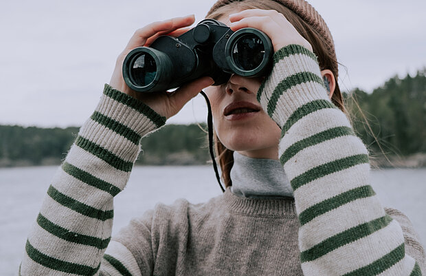 Frau mit gestreiftem Pullover und Schal, die mit einem Fernglas auf einen See blickt. Im Hintergrund sind Bäume und ein bewölkter Himmel zu sehen.