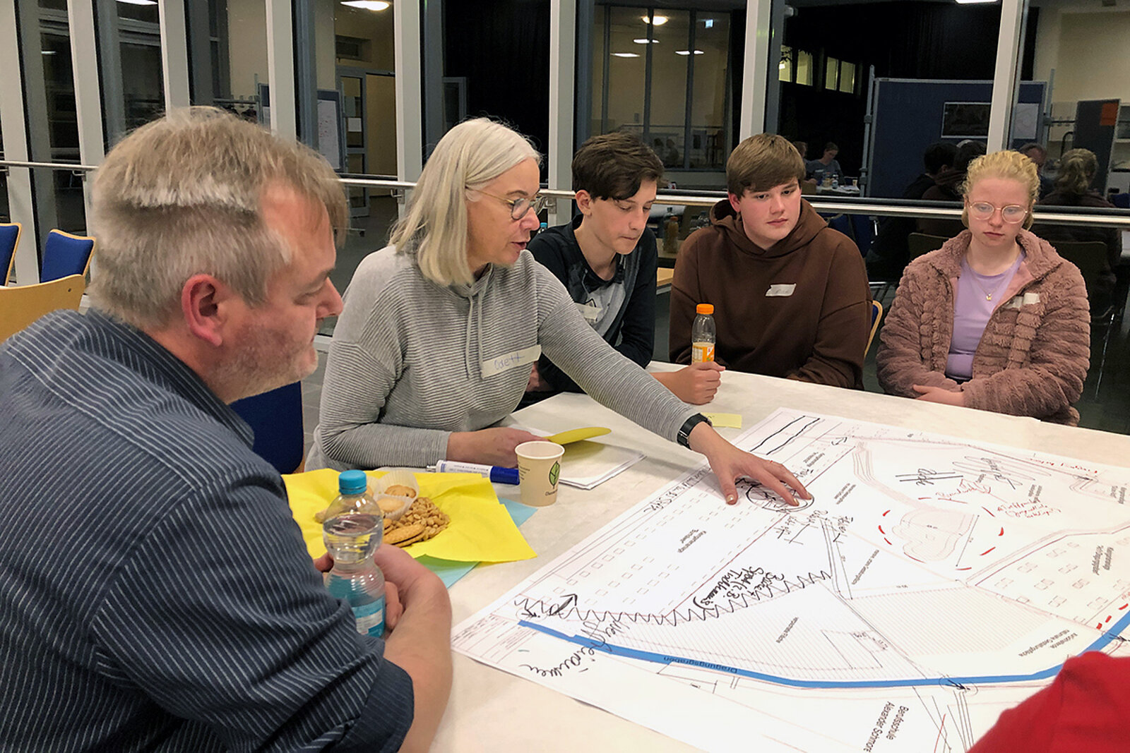 Gruppe von fünf Personen sitzen an einem Tisch und schauen auf einen großen ausgebreiteten Plan auf dem Tisch.