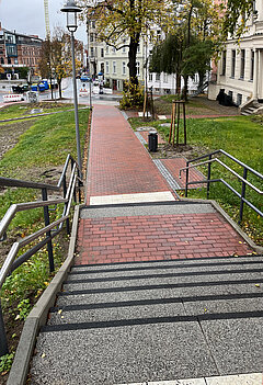 Blick von einer Treppe auf einen roten Backsteinweg, der durch eine Grünfläche zu einer Straße mit Häusern führt
