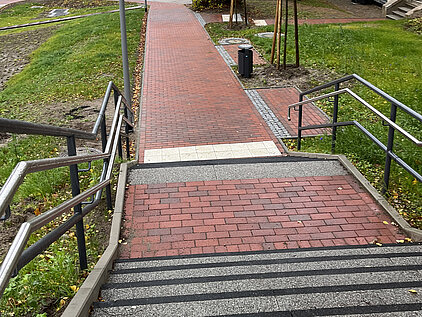 Blick von einer Treppe auf einen roten Backsteinweg, der durch eine Grünfläche zu einer Straße mit Häusern führt