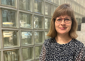 Frau mit Brille und bluse mit Tiermuster, lächelt vor einer Wand aus Glasbausteinen.