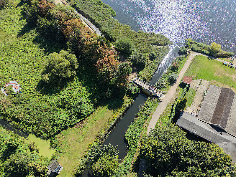 Luftaufnahme eines schmalen Wasserkanals mit Brücke, umgeben von Bäumen, Wiesen und Gebäuden neben einem größeren Gewässer