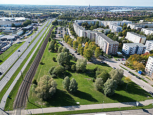 Luftaufnahme eines Wohngebiets mit mehrstöckigen Häusern, angrenzendem Park mit Bäumen und Bahngleisen links