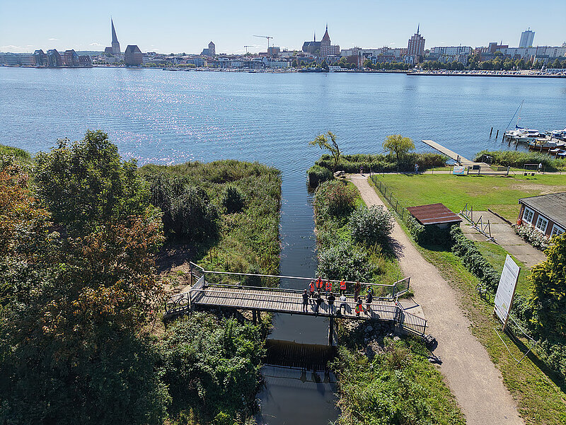 Blick von oben auf eine schmale Brücke über einen Wassergraben mit mehreren Personen darauf, im Hintergrund die Warnow und Rostocks Silhouette mit hohen Gebäuden
