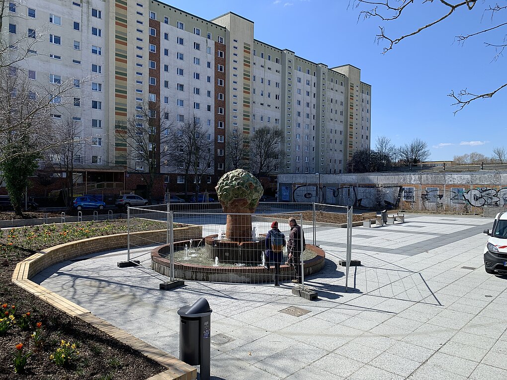 Baustelle mit mehreren Arbeitern und Baumaschinen vor mehrstöckigen Wohngebäuden unter klarem Himmel.