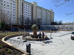 Baustelle mit mehreren Arbeitern und Baumaschinen vor mehrstöckigen Wohngebäuden unter klarem Himmel.