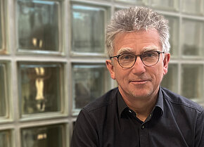 Mann mit grauen Haaren und Brille, trägt ein schwarzes Hemd, sitzt vor einer Wand aus Glasbausteinen.