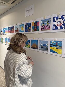 Frau mit hellem, lockigem Haar und weißem Strickpullover betrachtet bunte Bilder an einer weißen Wand.
