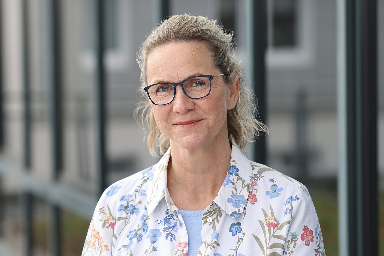 Frau mit hochgestecktem blonden Haar trägt eine weiße Bluse mit floralem Muster vor unscharfem Hintergrund eines Gebäudes