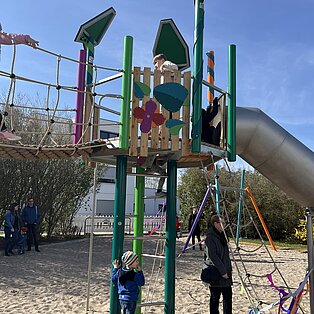 Kinder spielen auf einem grünen Klettergerüst mit Rutsche und Sandkasten bei sonnigem Wetter