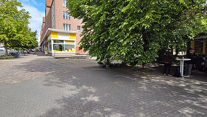 Straßenecke mit einem gelb-weißen Laden und schattigem Bereich mit Bäumen und zwei Personen an Stehtischen