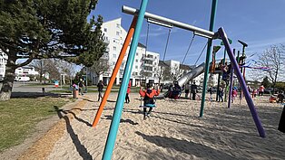 Kind schaukelt auf einer bunten Schaukel im Sand auf einem Spielplatz mit Rutschen und Klettergeräten, umgeben von Wohngebäuden