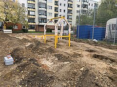 Baustelle mit Erde und Baugeräten vor einem Wohngebäude mit Balkonen.