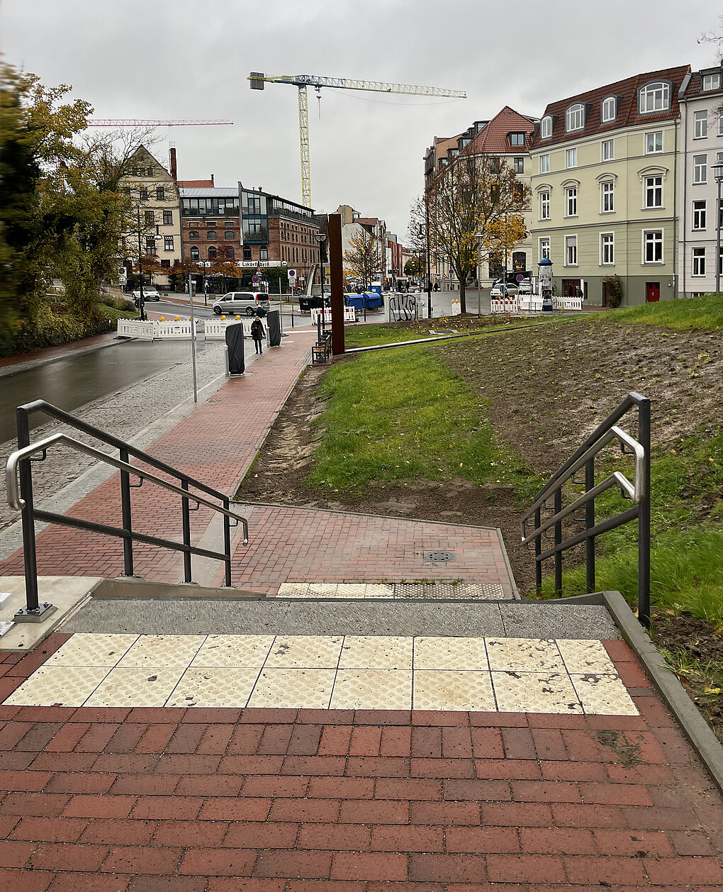 Blick von einer Treppe mit taktilen Bodenplatten auf eine Straße mit Baustellenabsperrungen, Bäumen und mehrstöckigen Wohnhäusern im Hintergrund