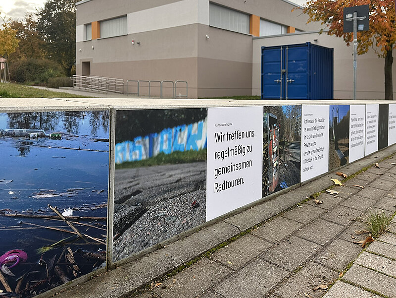 Sitzbank mit Fotomotiven und Texttafeln entlang eines Bürgersteigs vor einem Gebäude und einem blauen Container