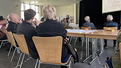 Menschen sitzen in einem hellen Raum auf Stühlen und schauen auf eine Leinwand mit einer Präsentation